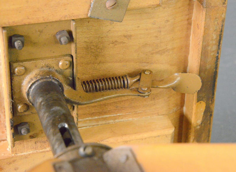 Op de afbeelding: Close-up van een houten tafel met een metalen veermechanisme. Het mechanisme heeft een metalen staaf en een hendel met een plat, rechthoekig pedaal. De tafel is gemaakt van licht hout en heeft een natuurlijke afwerking.