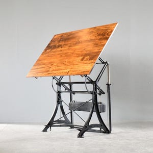 May include: A drafting table with a large, rectangular, polished wooden top set at an angle. The table is supported by a heavy-duty, black cast iron base featuring an intricate mechanical frame with adjustable levers, metal rods, and a central weight.