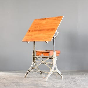 May include: A vintage drafting table featuring a large, rectangular, light brown wooden surface tilted at an angle. The table is supported by a sturdy, off-white metal base with a mechanical adjustment mechanism and a wooden storage drawer underneath.