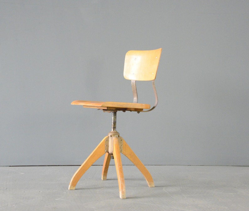 Op de afbeelding: Een vintage draaibare houten stoel met een metalen frame en een lichte houtkleur. De stoel heeft een eenvoudig ontwerp met een afgeronde rugleuning en een vierpotig onderstel.