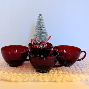 Puede incluir: Cuatro tazas de té de vidrio rojo rubí con exteriores texturizados están dispuestas sobre una superficie blanca texturizada. Un pequeño árbol de Navidad decorativo y un cuenco con una cinta roja y verde están en el fondo.