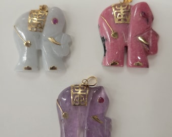 May include: Three carved elephant pendants with gold accents. The elephants are made of white, pink, and purple stone. Each elephant has a small red gemstone for an eye.