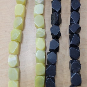 May include: Three strands of beads. Two strands are black, faceted, and geometric. The third strand is composed of light green, cube-shaped beads. The beads are strung on a thin, light-colored string. The beads are displayed on a light brown surface.