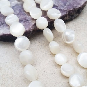 May include: A strand of white mother-of-pearl beads, each bead is round and flat, with a slightly iridescent sheen. The beads are strung on a clear thread and are lying on a white surface.