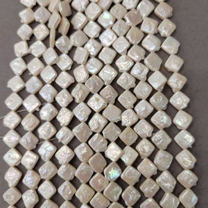 May include: A strand of white iridescent square-shaped pearl beads. The beads are strung on a white thread and have a slightly irregular shape.