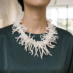 May include: A statement necklace composed of numerous white, branch-like structures that mimic the appearance of coral. The pieces are densely clustered together to form a wide, textured collar that rests around the neck, contrasting against a dark green top.
