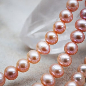 May include: A strand of pink pearls on a white background. The pearls are round and smooth, and they are strung together on a thin thread.