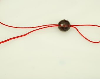 Könnte beinhalten: Eine facettierte, dunkelrote Perle auf einer leuchtend roten Kordel. Die Kordel ist geschlungen und bildet ein einfaches Design. Die Perle hat ein geometrisches Muster, und die Kordel ist dünn und flexibel.