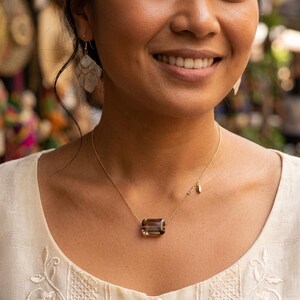 May include: A gold chain necklace featuring a rectangular, emerald-cut smoky quartz pendant. The necklace includes a small, delicate gold charm attached to the chain near the pendant. The jewelry is worn against a cream-colored textured top.