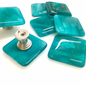 Op de afbeelding: Set van zes vierkante teal glazen kastknoppen met zilveren metalen bases. De knoppen hebben een doorschijnende, licht troebele uitstraling.
