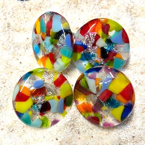 May include: Four colorful glass cabochons with a mosaic design. The cabochons are oval and round, featuring a mix of red, yellow, blue, and green glass pieces. Some have silver foil accents, set against a sandy background.
