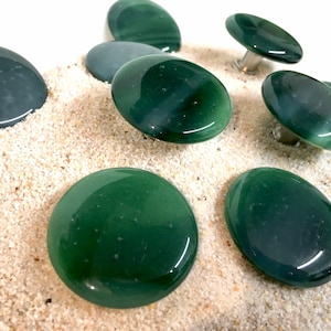 Peut inclure: Un ensemble de sept boutons ronds en verre vert avec une finition lisse et brillante. Les boutons sont disposés sur une surface de sable blanc.