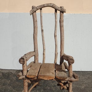 May include: A rustic wooden chair constructed from natural branches and planks. The chair features a high back with vertical supports and armrests made from curved branches. The seat is composed of three wooden planks. The chair's natural wood tones create a unique, organic aesthetic.