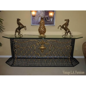 May include: A black wrought iron console table with a glass top and gold accents. The table has a decorative design with a central motif and a diamond pattern. Two gold horse sculptures are on the table, and a gold pineapple-shaped lamp is in the background.