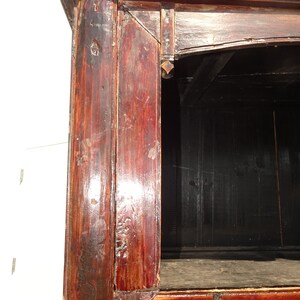 Op de afbeelding: Close-up van de hoek van een donkerrode houten kast met een zwart interieur. Het hout heeft een verweerde afwerking en vertoont tekenen van ouderdom.