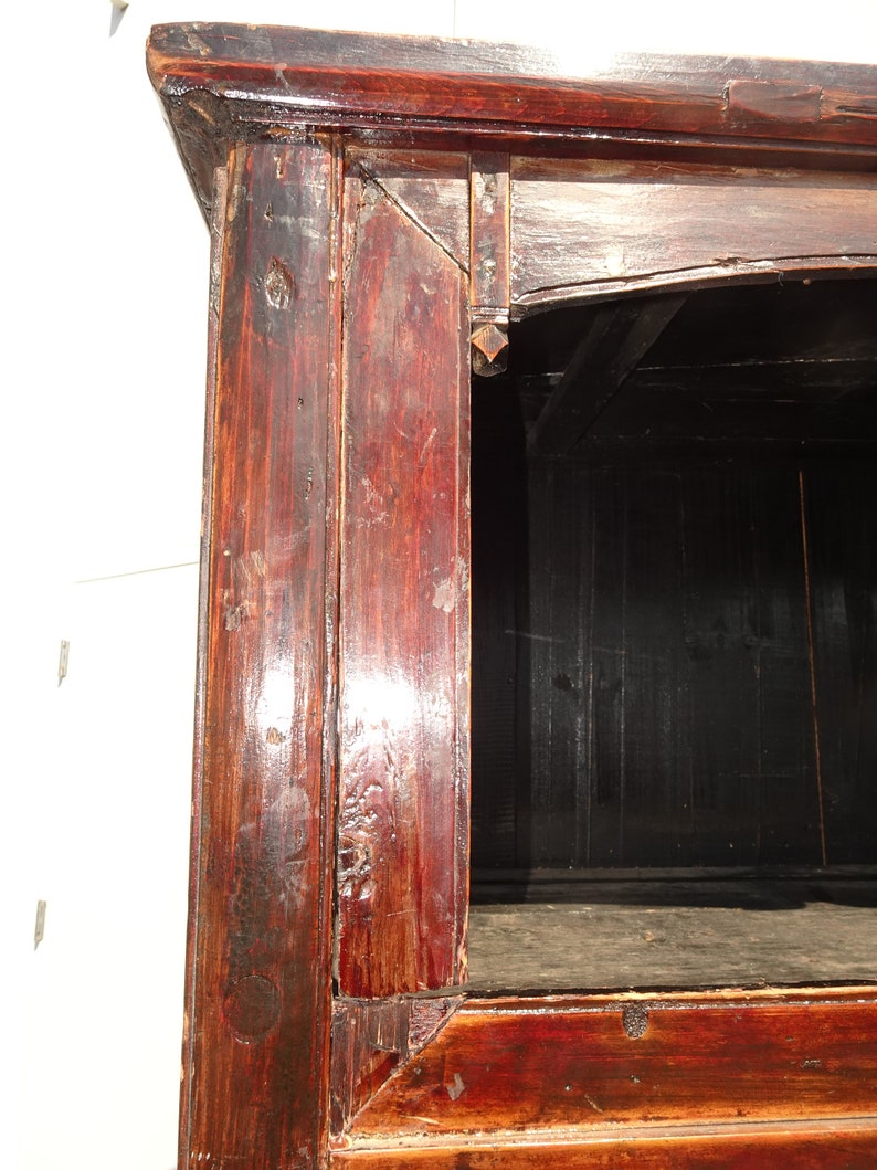 Op de afbeelding: Close-up van de hoek van een donkerrode houten kast met een zwart interieur. Het hout heeft een verweerde afwerking en vertoont tekenen van ouderdom.