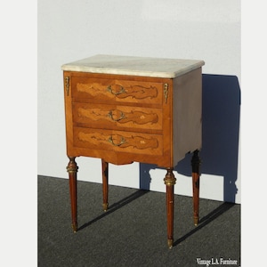 May include: A three-drawer nightstand with a white marble top and intricate marquetry details. The nightstand has four slender legs and brass hardware.