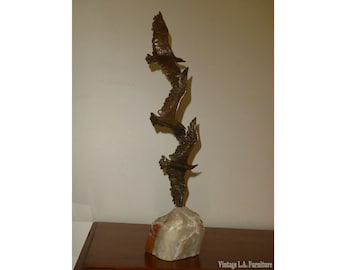 May include: A bronze sculpture of three birds in flight, mounted on a white marble base. The birds are arranged in a vertical line, with the top bird's wings spread wide. The sculpture is a modern design and would make a great addition to any home.