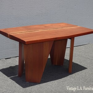May include: A solid wood coffee table with a unique, angular design. The table top is made of a single piece of wood with a natural grain pattern. The legs are also made of wood and have a geometric shape.