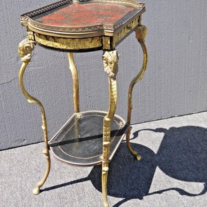 May include: A two-tiered side table with a black and red marbled top and gold-painted legs. The legs are ornately decorated with a floral design and have a ram's head at the top.