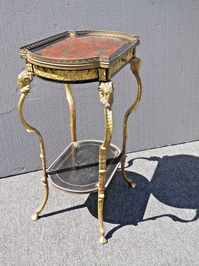 May include: A two-tiered side table with a black and red marbled top and gold-painted legs. The legs are ornately decorated with a floral design and have a ram's head at the top.