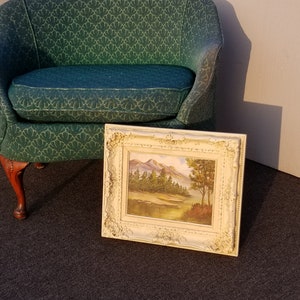 May include: A framed landscape painting of a mountain scene with trees and a river. The painting is in a white frame with ornate details. The painting is sitting on a dark carpet in front of a green chair.