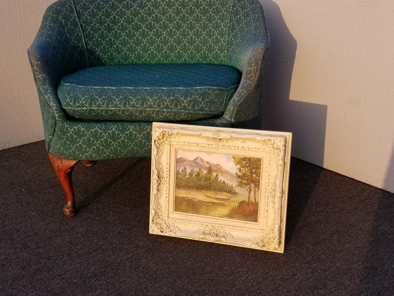 May include: A framed landscape painting of a mountain scene with trees and a river. The painting is in a white frame with ornate details. The painting is sitting on a dark carpet in front of a green chair.