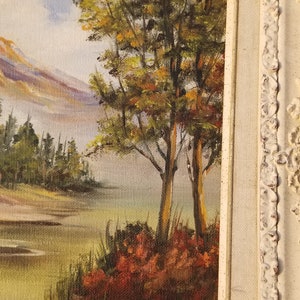 May include: A framed oil painting depicting a landscape scene with a mountain range in the background, a lake in the foreground, and a group of trees in the center. The trees are painted in shades of green, brown, and yellow, and the lake is a light blue color. The mountain range is painted in shades of purple, blue, and white.