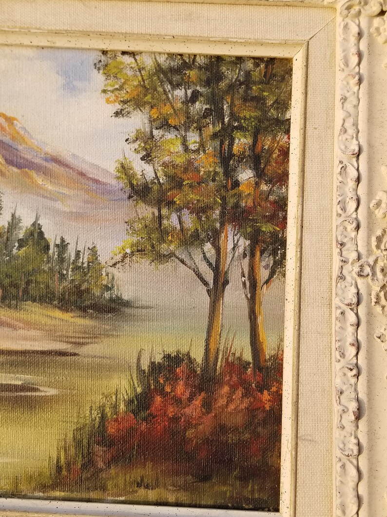 May include: A framed oil painting depicting a landscape scene with a mountain range in the background, a lake in the foreground, and a group of trees in the center. The trees are painted in shades of green, brown, and yellow, and the lake is a light blue color. The mountain range is painted in shades of purple, blue, and white.