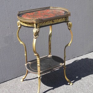 May include: A two-tiered side table with a black and gold finish. The top tier has a red and gold marbled surface with a gold trim. The bottom tier is black with a gold trim. The table has four legs with gold ram's head accents.