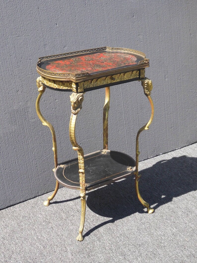 May include: A two-tiered side table with a black and gold finish. The top tier has a red and gold marbled surface with a gold trim. The bottom tier is black with a gold trim. The table has four legs with gold ram's head accents.