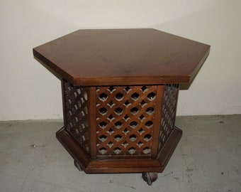 May include: A dark brown, hexagonal side table with a decorative lattice design on the sides. The table has a solid top and sits on small wheels. The table is made of wood.