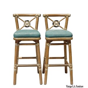 May include: Two bamboo bar stools with teal seat cushions. The backs feature an X-shaped design with a decorative center. The stools have a natural wood finish and are tied together with rope detailing. Vintage L.A. Furniture.