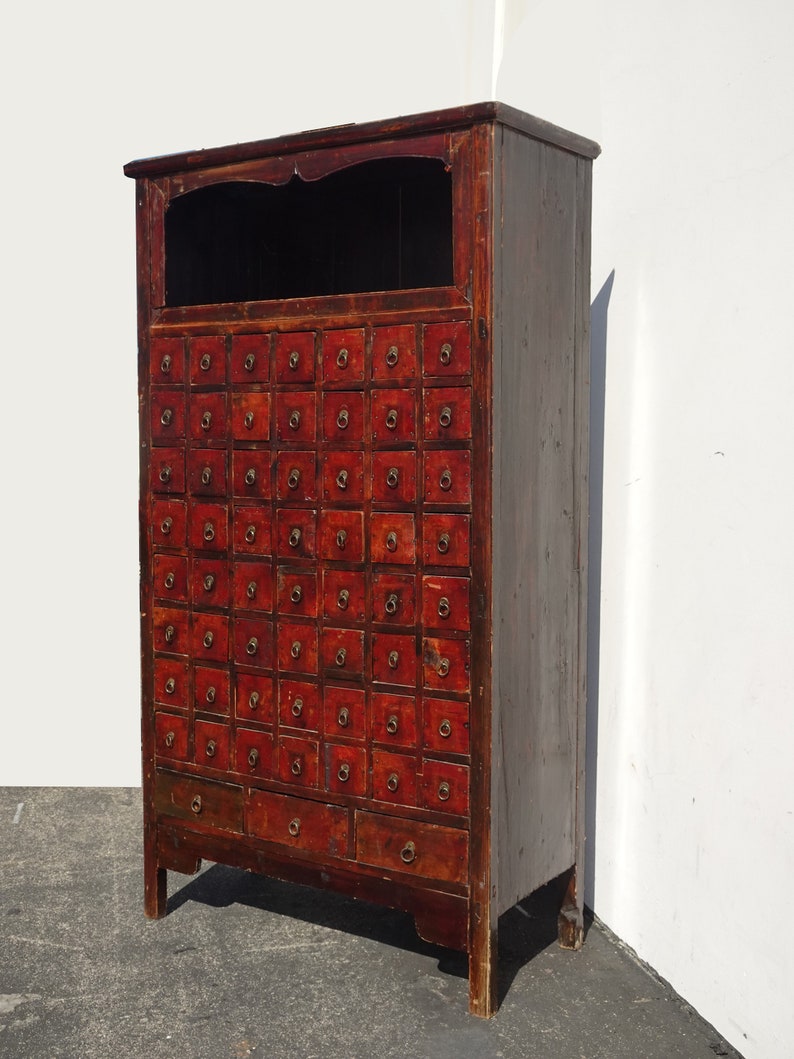 Op de afbeelding: Een hoge, antieke, rode houten kast met een reeks kleine laden. De kast heeft een bovenste plank en een paar grotere laden aan de onderkant.