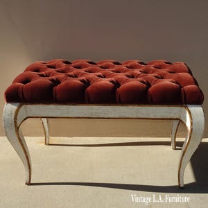 May include: A white painted wooden bench with a tufted red velvet seat. The bench has four legs with a curved design.