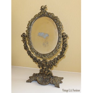 May include: A vintage gold-toned metal vanity mirror with an oval-shaped glass and ornate floral and cherub detailing. The mirror has a swivel stand with a decorative base.