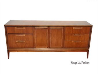 May include: A long wooden dresser with a smooth top surface, featuring three drawers on the left side, three drawers on the right side, and two central cabinet doors. Each drawer and door has a horizontal handle. Text reads Vintage L.A. Furniture.