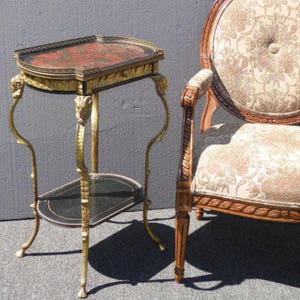 May include: A gold and black two-tiered side table with a decorative top and ornate legs. The table is next to a chair with a floral patterned fabric upholstery.