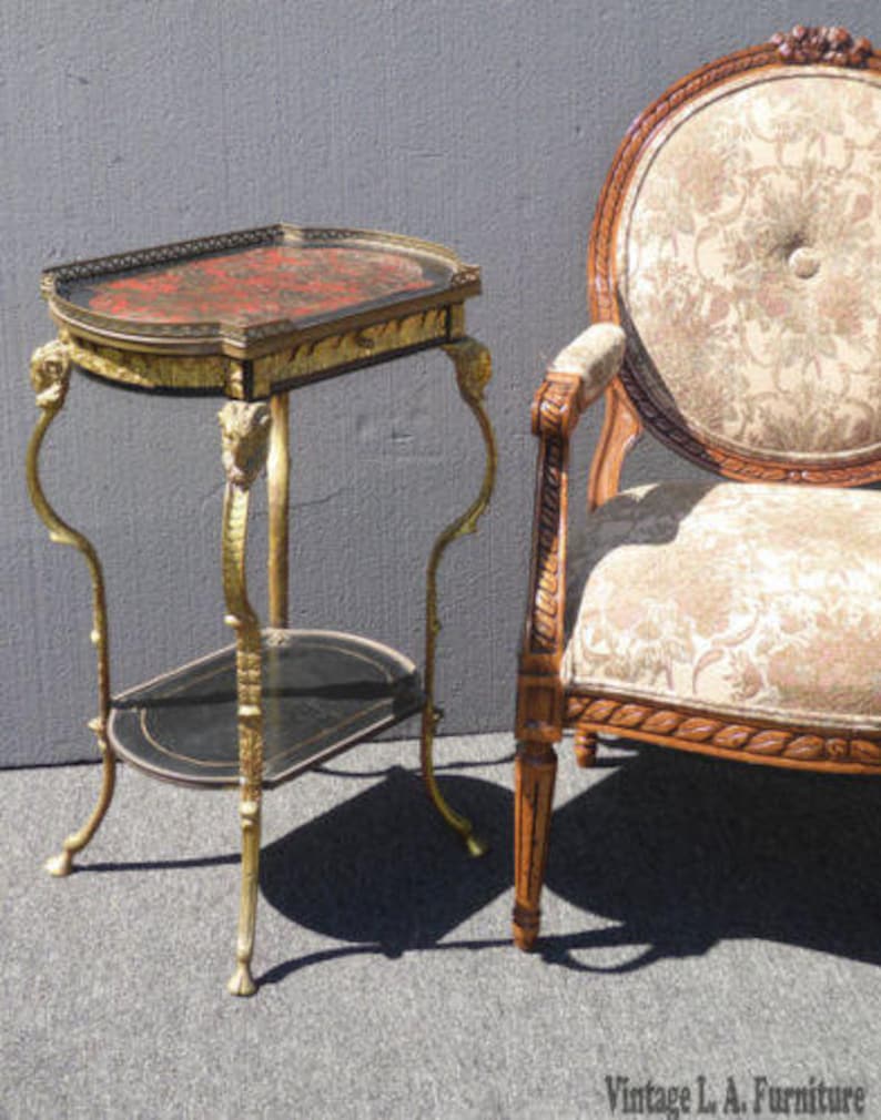 May include: A gold and black two-tiered side table with a decorative top and ornate legs. The table is next to a chair with a floral patterned fabric upholstery.