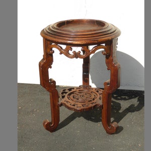 May include: A dark brown wooden plant stand with intricate carvings and a round top. The stand has three legs with curved feet.