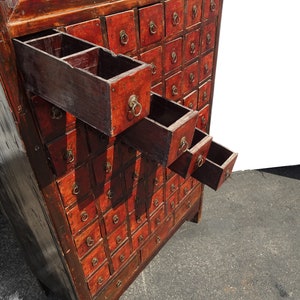 Op de afbeelding: Antiek houten kast met meerdere kleine laden, elk met een messing ringgreep. De laden zijn rood geverfd en hebben een verweerde afwerking. Sommige laden zijn open, waardoor het interieur zichtbaar is.