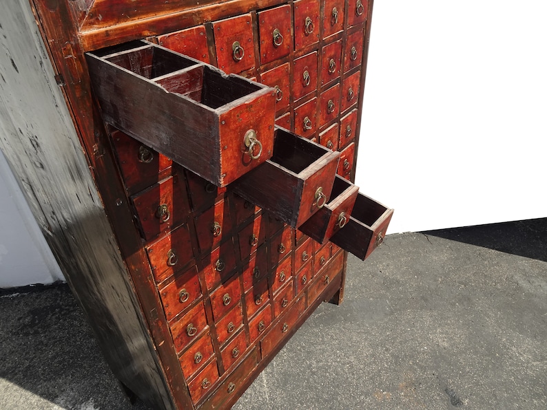 Op de afbeelding: Antiek houten kast met meerdere kleine laden, elk met een messing ringgreep. De laden zijn rood geverfd en hebben een verweerde afwerking. Sommige laden zijn open, waardoor het interieur zichtbaar is.