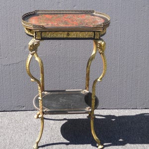 May include: A gold-toned metal side table with a black and red marquetry top. The table has a lower shelf and ornate legs.