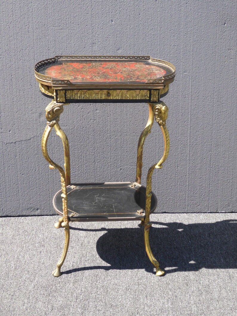 May include: A gold-toned metal side table with a black and red marquetry top. The table has a lower shelf and ornate legs.