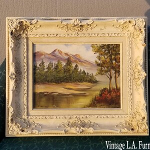 May include: A framed landscape painting depicting a lake with trees and mountains in the background. The frame is white with ornate details. The painting is signed "D. Warner".