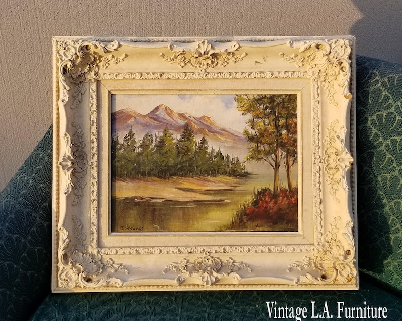 May include: A framed landscape painting depicting a lake with trees and mountains in the background. The frame is white with ornate details. The painting is signed "D. Warner".