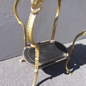 May include: A gold-painted metal table with two black shelves. The table has ornate, sculpted legs with ram's heads at the top.