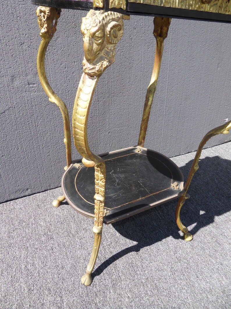 May include: A gold-painted metal table with two black shelves. The table has ornate, sculpted legs with ram's heads at the top.