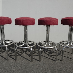 May include: Four bar stools with burgundy patterned seats and chrome-colored metal frames. The stools have a circular footrest and a curved base. The seats are round and padded.
