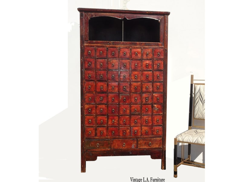 Op de afbeelding: Een vintage rode Chinese apothekerskast met een donkerhouten bovenblad en veel kleine laden. De kast heeft een verweerde uitstraling en is gemaakt van hout.